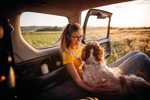 Woman sits in the trunk of a car with her dog and watches the sunset